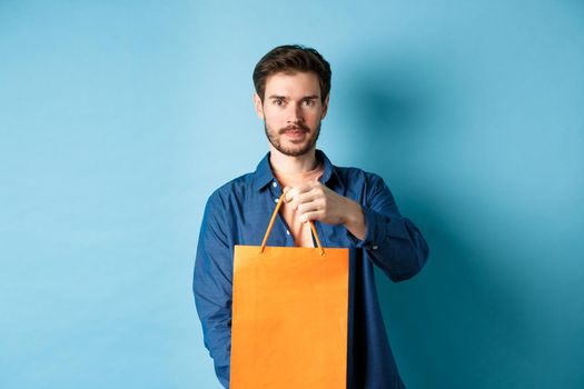Handsome bearded man stretch out hand with orange shopping bag, making a gift, standing on blue background.