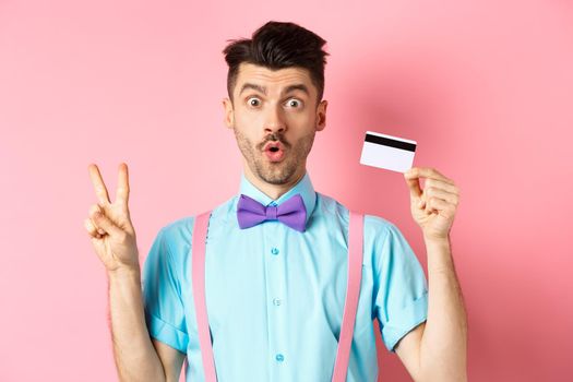 Shopping concept. Image of funny guy looking surprised while showing plastic credit card and victory sign, checking out promo deal, standing on pink background.