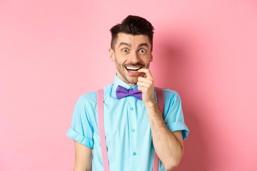 Excited and tempted young guy wants something, biting fingernail with temptation looking at camera, choosing product, standing over pink background.