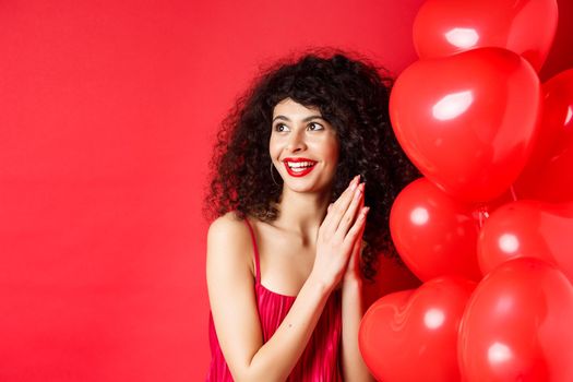 Excited beautiful woman with curly hair, standing near heart balloons and rubbing palms together, expect good deal or relish, red background.