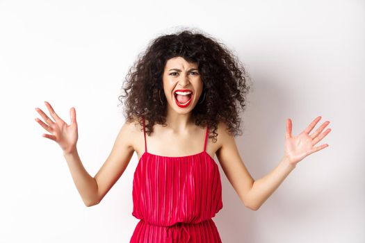 Angry young woman with curly hair, wearing red dress, screaming and having an argument, looking with hatred and anger, standing over white background.