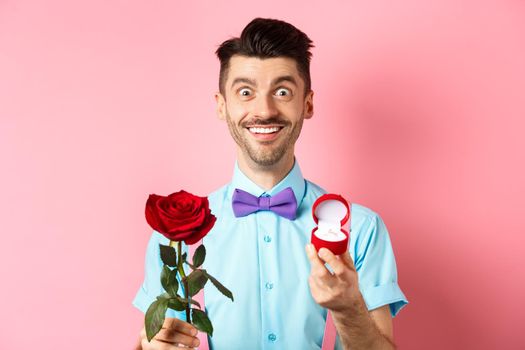 Valentines day. Smiling handsome man asking to marry him, showing engagement ring and red rose, standing romantic on pink background.