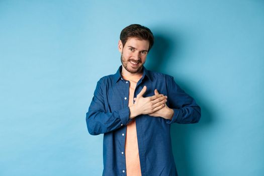 Smiling man feeling touched and grateful, holding hands on heart and thanking, standing on blue background. Copy space