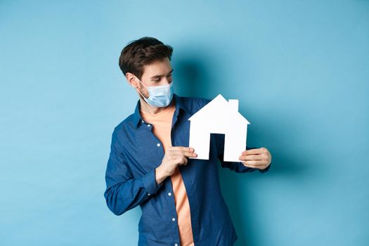 Real estate and quarantine concept. Young caucasian man in medical mask looking curious at paper house cutout, standing on blue background.