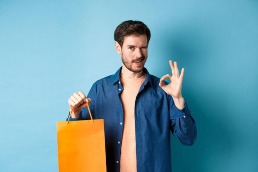 Modern man in casual outfit showing shopping bag and okay sign, winking at camera, recommending shop, standing on blue background.