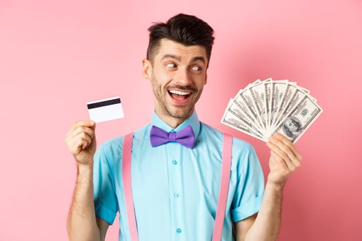 Image of smiling young man in bow-tie holding plastic credit card and looking happy at money, choosing cash, standing over pink background.