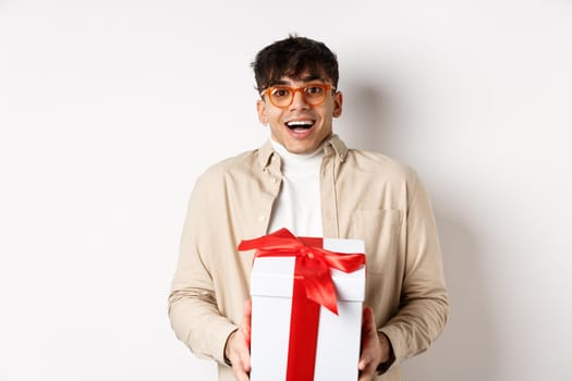 Surprised happy guy receiving a gift, holding present box and smiling amazed, standing on white background.