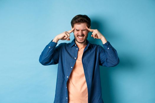 Man feeling sick, touching head and grimacing from painful migraine, having headache or feel dizzy, standing on blue background.