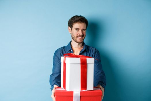 Handsome boyfriend winking at camera and wishing happy valentines day, extending hands with gift boxes, making romantic surprise, standing over blue background.