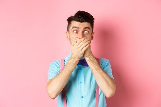 Funny young man covering mouth with hands and squint eyes, making clown grimaces, standing silly on pink background, entertain people.