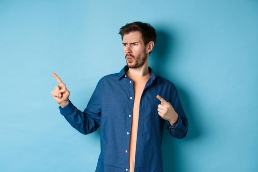 Confused and annoyed guy pointing and looking left at empty space with angry frowning face, standing on blue background.