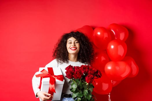 Smiling happy woman holding box with gift and red roses from boyfriend, celebrating Valentines day, standing near romantic hearts balloons, standing over studio background.