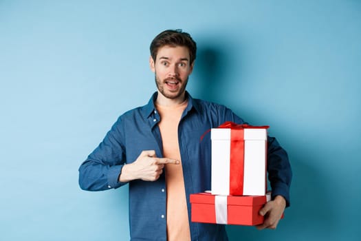 Handsome young man pointing at valentines gift boxes, standing over blue background in casual clothes. Copy space