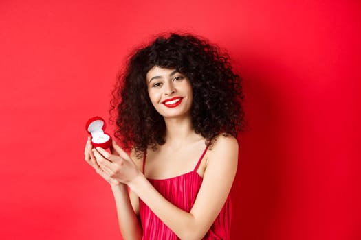 Romantic woman with curly hair, wearing red dress and showing engagement ring, standing happy on studio background.