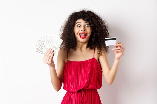 Happy attractive woman in red dress, screaming of joy and showing plastic credit card with money, winning prize, standing over white background.