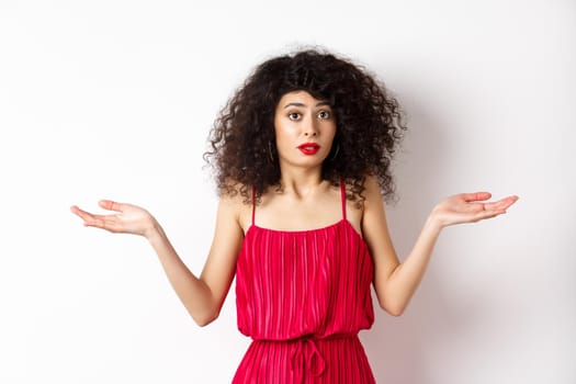 Confused lady in red dress shrugging shoulders, spread hands sideways and looking puzzled, know nothing, cant understand, standing over white background.