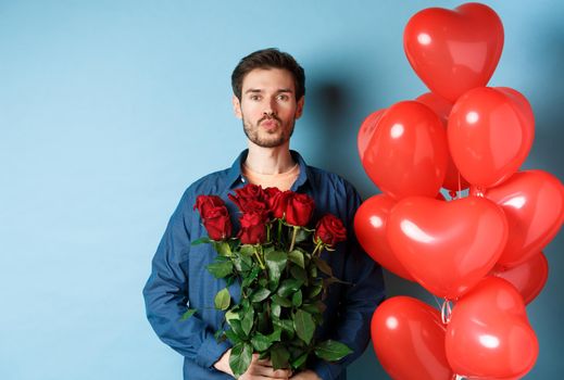 Romantic man with red roses and red heart balloons, pucker lips for kiss, making surprise on Valentines day, standing against blue background.