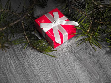 Close up photograph of a red small gift wrapped in white bow laying on a gray wood grain floor with evergreen pine needle limb in background.