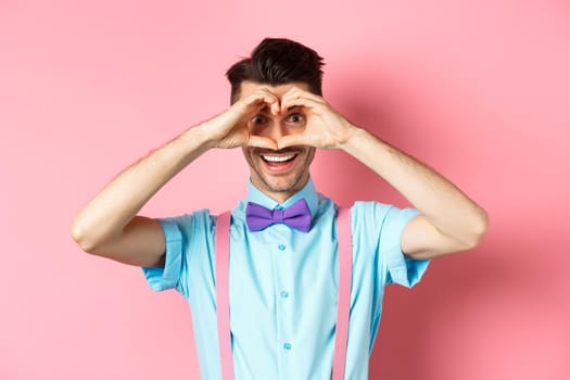 Happy Valentines day. Young man making hand heart and looking through it at camera, feeling romantic with lover, standing over pink background.