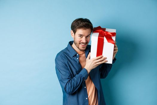 Valentines day. Romantic boyfriend showing gift box for lover, celebrating anniversary, standing over blue background.