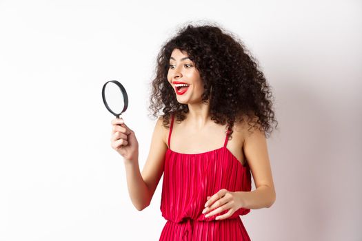 Excited woman in red dress look through magnifying glass and smiling, found interesting promo, standing on white background.