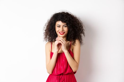 Cunning young woman with curly hair, steeple fingers and smiling devious, having a plan, standing in red dress on white background.