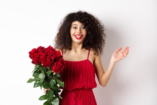 Romance and Valentines day. Woman gasping surprised and happy, receive surprise gift from lover, holding bouquet of red roses, standing on white background.