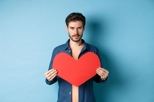 Young man looking for soulmate on valentines day, holding big red heart and looking at camera, standing over blue background.