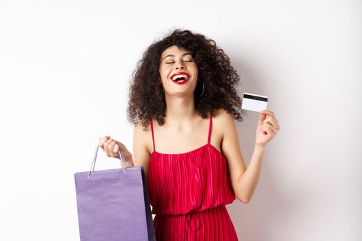 Happy woman in red dress, laughing and shopping, holding plastic credit card and store bag, standing on white background.