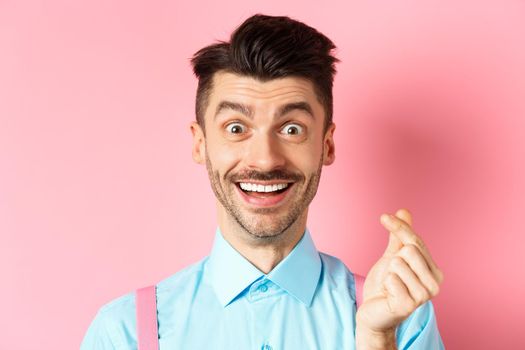 Valentines day concept. Smiling man showing finger heart and looking happy, standing over pink background.