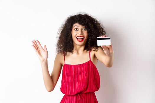 Shopping. Amazed curly-haired woman in red dress, showing plastic credit card and scream from excitement, standing on white background.