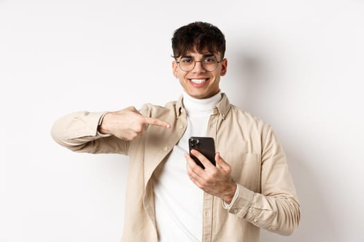 Real people. Handsome young man in glasses pointing finger at smartphone screen, showing online promo, standing on white background.