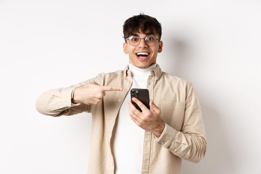 Technology and online shopping concept. Happy hipster guy pointing finger at smartphone screen and smiling, checking out app promo, standing on white background.