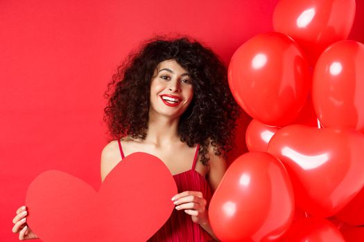 Happy valentines day. Romantic girl in dress waiting for true love, showing big red heart and standing near balloons on red background.