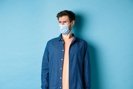 Covid-19 and healthcare concept. Dissatisfied frowning guy in medical mask looking at bad promo, stare aside at empty space with disappointed face, blue background.
