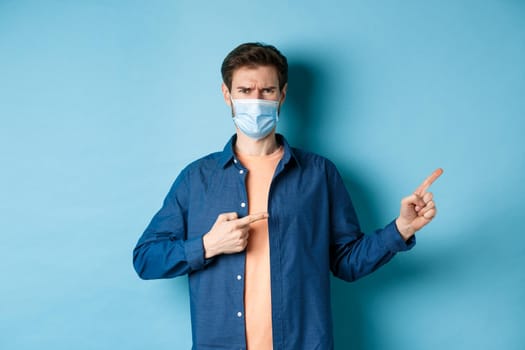 Covid-19 and pandemic concept. Angry man in medical mask frowning, pointig fingers at upper right corner empty space, complaining, standing on blue background.