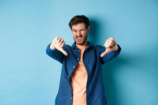 Disgusted guy express negative opinion, showing thumbs down and tongue, frowning upset, disapprove something bad, standing on blue background.