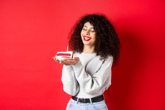 Happy birthday girl celebrating and making wish, holding bday cake and smiling, standing on red background.