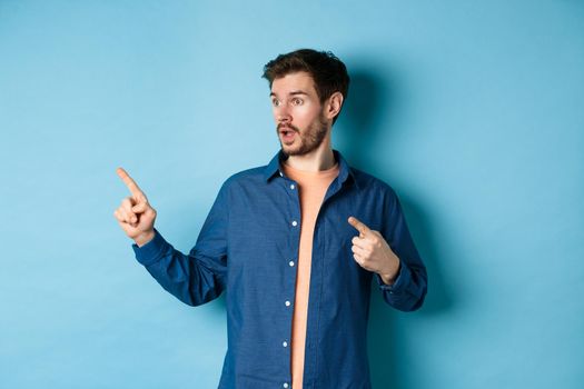 Surprised guy in casual clothes, pointing and looking left at empty space, showing advertisement, standing on blue background.