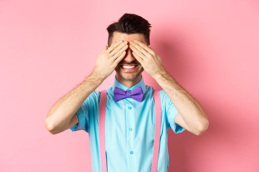 Image of smiling handsome guy waiting for surprise with closed eyes, standing in fancy outfit on pink background.
