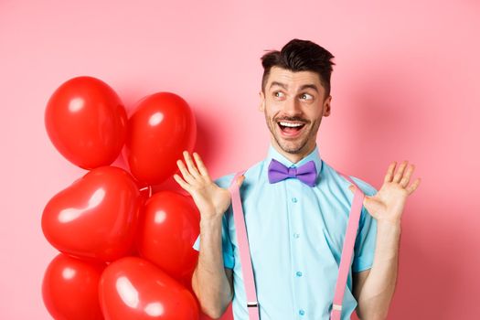 Love and romance concept. Happy man screaming from fantastic news, shouting wow and smiling amused, checking out special offer on Valentines, standing near red hearts balloons.