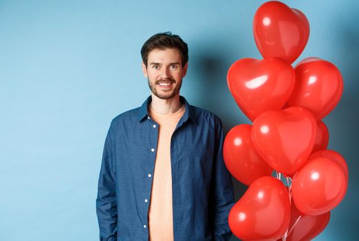 Cute boyfriend looking happy and smiling, standing near Valentines day heart balloons on blue background.