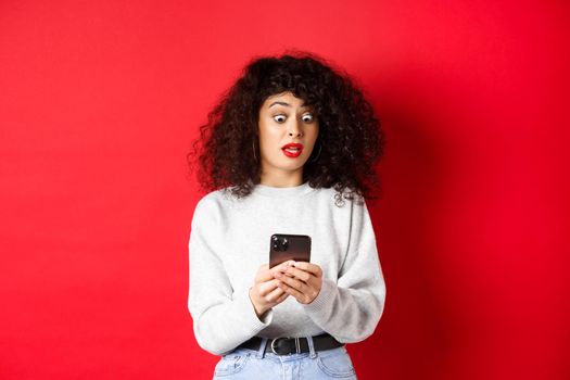 Shocked woman stare at smartphone screen with popped eyes, reading strange message, standing on red background.