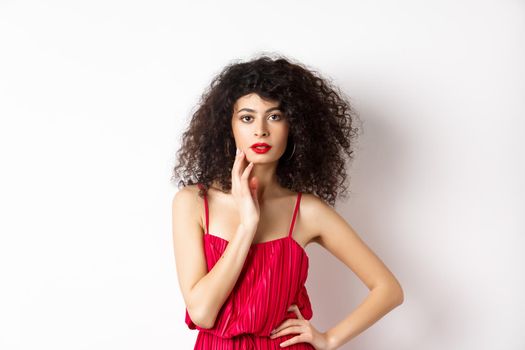 Beauty and fashion. Sensual female model with curly hair and red lips, wearing elegant dress, touching face and looking seductive, white background.