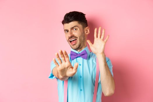 Silly young man in bow-tie and suspenders asking to stop, covering face with hands and screaming terrified, looking at something disgusting, standing on pink background.