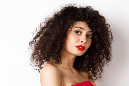 Close-up of beautiful caucasian woman with curly haircut and red lipstick, looking tender at camera, standing over white background.