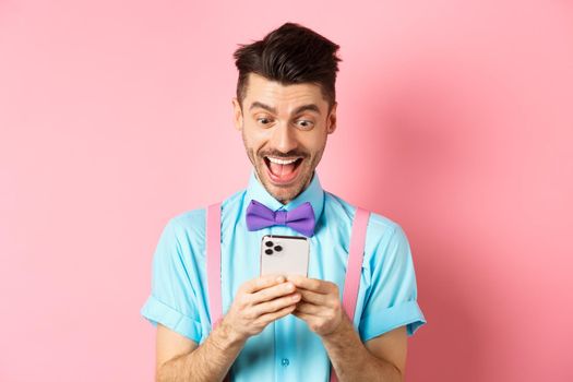 Technology concept. Cheerful young guy winning online prize, staring happy at smartphone screen, reading message amazed, standing on pink background.