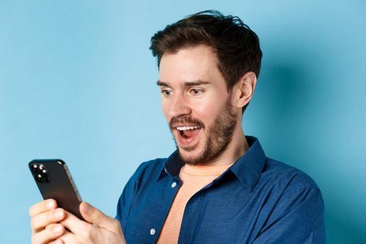 Image of handsome young man feeling happy reading good news on mobile phone, standing cheerful against blue background.