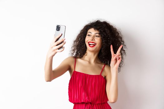 Elegant young woman in red dress taking selfie on smartphone, posing on event party, standing with mobile phone on white background.