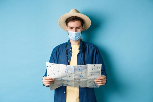 Covid-19, pandemic and travel concept. Young man in medical mask reading tourist map, searching for route to sightseeings, standing on blue background.